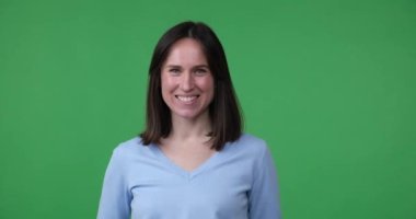 Güzel, beyaz bir kadın yeşil bir ekranda kendine güvenerek duruyor. Gülümsüyor, kolları göğsünde çapraz duruyor. Kendine güvenen tavrını ve pozitif enerjisini gösteriyor..