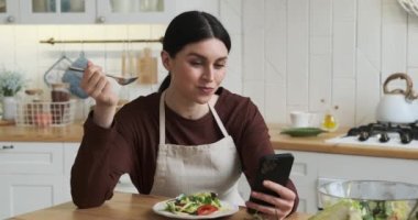 Beyaz bir kadın mutfak masasında oturmuş tabağındaki salatanın tadını çıkarırken aynı anda telefonuyla meşgul oluyor. Geniş bir gülümseme ve bulaşıcı bir kahkaha yayıyor..
