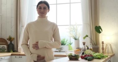 Işıl ışıl hamile bir kadın mutfak masasının yanında duruyor ve kameraya yumuşak bir gülümsemeyle bakıyor. Eli sevgi dolu bir şekilde yuvarlak karın üzerinde süzülür, anne ve çocuk arasındaki derin bağı ifade eder..