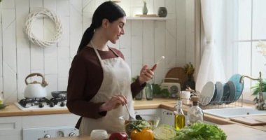 Mutfak önlüğü giymiş, kendinden emin, beyaz bir kadın. Cam bir kâse taze salatayla ahşap kaşıkla malzemeleri ustalıkla harmanlıyor. Karıştırırken, telefonunu kontrol ediyor..