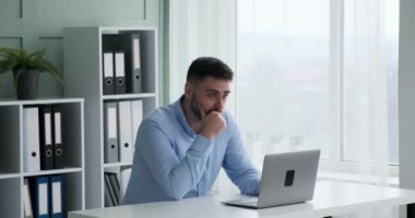 Ofisinde oturan, laptopunu rahatça ve odaklanmış bir şekilde yazan, kendinden emin beyaz iş adamı. Yüzünde hoş bir gülümsemeyle, işinden zevk alıyor gibi görünüyor..