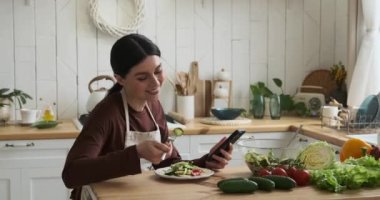 Neşeli Kafkasyalı kadın mutfak masasında taze salata yiyor. Her ısırığın tadını çıkararak telefonunu karıştırıyor, teknolojiyi ve yemeği harmanlıyor..
