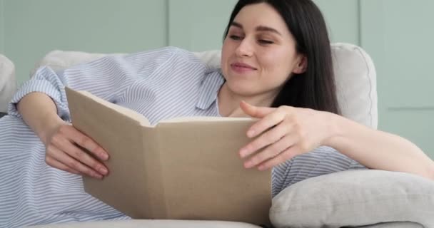 ソファに横になって本を読んでいる満足した白人女性 リラックスした笑顔と積極的なエネルギーで 彼女は読書の喜びに没頭しています — ストック動画