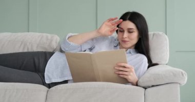 Kanepede uzanmış gülümseyen bir kitap okuyan mutlu bir beyaz kadının portresi. Rahatlamış ve mutlu ifadesi, okumanın ve sakin bir anın zevkini kendisine aktarıyor..