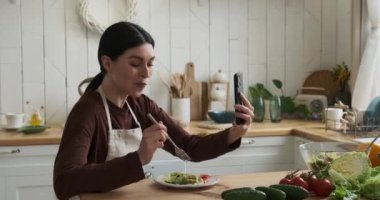 Mutfak masasındaki neşeli beyaz kadın lezzetli salatanın tadını çıkarıyor. Görüntülü arama yapıyor, sıcak bir gülümsemeyle parlıyor. Yemeği ve sanal etkileşimi dengelemek için çaba sarf etmeden pozitif enerji yayıyor..