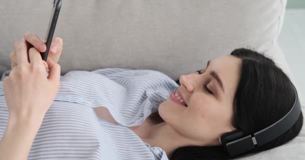 一个白人妇女躺在沙发上 通过无线耳机欣赏音乐 并愉快地在手机上发短信的画像 她脸上挂着笑容 在周围环境中很放松 — 图库视频影像