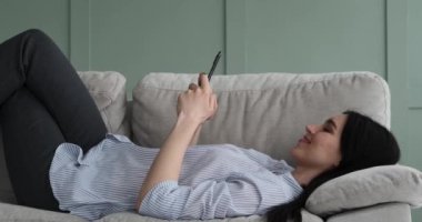 Mutlu ve neşeli beyaz kadın koltukta uzanıyor, cep telefonunda daktilo ve mesaj yazıyor. Yüzünde bir gülümseme var ve telefonda konuşurken ya da mesajlaşırken rahatlamış görünüyor..