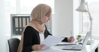 Takım elbiseli, beyaz gömlekli ve tesettürlü genç bir Arap iş kadını ofis masasında oturuyor, evrak işleriyle meşgul, bilgisayarında daktilo yazıyor. Not defterine not alıyorum..