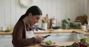 Neşeli beyaz kadın mutfak masasında oturuyor, tabaktan salata yiyor. Geniş bir gülümseme ile, telefonuyla canlı sohbete dalmışken pozitiflik ve mutluluk yayıyor..
