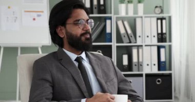 Takım elbiseli ve kravatlı Hintli işadamı rahat bir koltukta otururken, sıcak bir içecek yudumlarken ve yüzünde mutlu bir gülümsemeyle pencereden dışarı bakarken görülüyor..