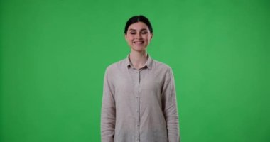 Yeşil arka planda duran beyaz bir kadın eliyle iyi bir hareket gösteriyor. Dostane bir ifadeyle doğrudan kameraya bakıyor ve kendinden emin ve pozitif görünüyor..