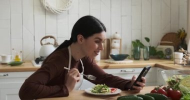 Neşeli bir Kafkasyalı kadın mutfak masasında oturmuş tabaktan salatanın tadını çıkarırken aynı anda telefonuna sarılmış. Işıl ışıl gülüşü ve bulaşıcı gülüşü neşe dolu tavırları.