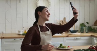 Neşeli beyaz kadın mutfak masasında oturmuş nefis salatanın tadını çıkarıyor. Video çağrısına dalmış, yemek ve sohbeti dengeleyerek sıcaklık ve pozitiflik yayıyor..