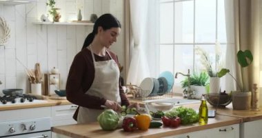 Beyaz kadın mutfak önlüğü lezzetli bir yemek hazırlar. Yeteneğiyle, tahta kesme tahtasındaki malzemeleri dilimliyor. Tahtayı kaldırarak, doğranmış malzemeleri cam bir kaseye dolduruyor..