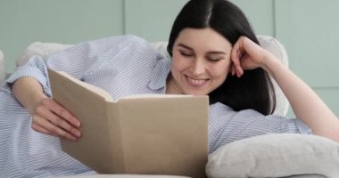 Kanepede uzanmış kitap okuyan neşeli beyaz bir kadının portresi. Gülümsüyor, okuduktan sonra gülüyor ve boş zamanlarının tadını çıkarıyor gibi görünüyor..