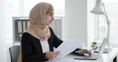 Ofis masasında oturan tesettürlü genç bir Arap iş kadını. Olumlu bir ruh hali içinde ve belgeleri inceliyor, bilgiyi bilgisayara giriyor ve klavyeyi ustalıkla çalıştırıyor..