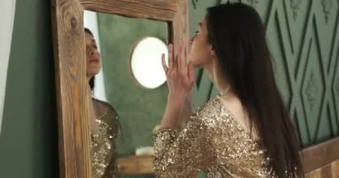 Beyaz kadın, gece elbisesi giymiş, kendini partiye hazırlıyor. Aynanın karşısında titizlikle saçını ve kusursuz makyajını ayarlar. Her detayın mükemmel bir uyum içinde olmasını sağlar..