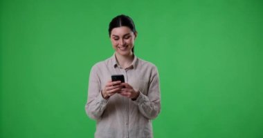Neşeli, beyaz bir kadın yeşil ekran arka planında cep telefonunu mesaj atmak ya da göz atmak için kullanırken duruyor. Çok fazla gülümsüyor, sonra mesaj atmaya devam ederken kahkahalara boğuluyor..