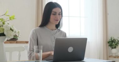 Mutfak masasında oturan odaklanmış beyaz bir kadın, dizüstü bilgisayarında yazarken işine dalmış. Üretken bir seanstan sonra, bir bardak suya uzanıyor, ferahlatıcı bir yudum ile susuzluğunu gideriyor..