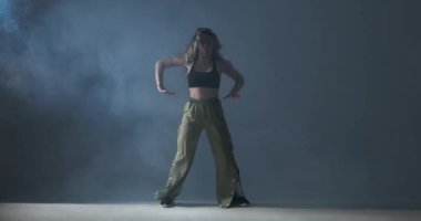 Kıvırcık saçlı, gri arka plan üzerinde beyaz bir kadın. İçi dumanla dolu. Dinamik dans figürlerinin sergilendiği bir gösteri. Canlı yeşil pantolonlar giyer, enerji, yaratıcılık ve tutku saçar..