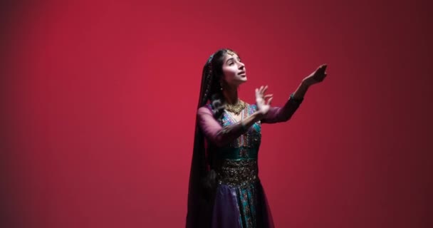 カラフルなサリーを着た若い女性としての伝統的なインドのダンスは 優雅に動き 鮮やかな赤を背景に彼女の動きを通して喜びを表現しています 彼女はインドの伝統的なジュエリーを着て — ストック動画