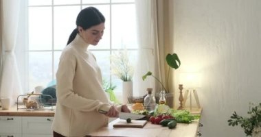 Hamile beyaz kadın mutfak tezgahının yanında duruyor. Son derece özen ve sevgiyle, ferahlatıcı bir salata hazırlar, lezzetli bir salatalığı ince ince doğrar ve canlı bir kaseye koyar..