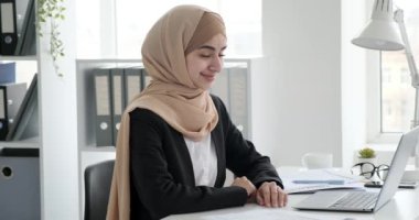 Ofiste masa başında oturan kendine güvenen bir Arap iş kadını. O bir tesettür, siyah bir ceket ve beyaz bir gömlek giyiyor. Özenli bir bakışla doğrudan kameraya bakar ve geniş bir şekilde gülümser..