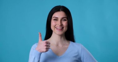 Beyaz ırktan neşeli bir kadın mavi arka planda duruyor, kameraya bakıyor ve yüzünde kocaman bir gülümsemeyle baş parmak işareti gösteriyor. Bir şeyi onaylıyor ve seviyor..