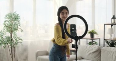 Oturma odasındaki genç Hintli kız sosyal medya için bir video kaydediyor ya da cep telefonu ve halka lambasıyla canlı yayına çıkıyor. Yüzünde geniş bir gülümsemeyle dans yeteneklerini sergiliyor..