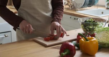 Mutfak önlüğü giymiş zarif bir beyaz kadın salata için domatesleri ustalıkla dilimliyor. Domates dilimlerini büyük bir dikkatle bir kaseye koyar..