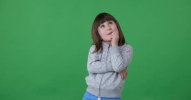 Sevimli anaokulu kızı duruyor, düşünüyor ve etrafı inceliyor. Gülümsüyor, becerikliliğini ve merakını gösteriyor ve etrafındaki her şeye ilgi ve ilgiyle bakıyor.