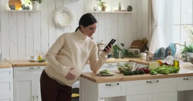 Mutfakta beyaz hamile bir kadının gözleri neşeyle doluydu. Telefon elindeyken, etrafı kolaçan ediyor. Diğer eli yavaşça karnını okşar, içinde büyüyen değerli hayata değer verir..