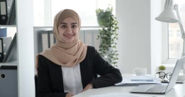 Genç bir Arap iş kadını, tesettüre bürünmüş, pozitiflik ve güven saçıyor. Yüzünde geniş bir gülümsemeyle, ofisteki masasına oturur, neşeli ve canlı bir ruh hali yayar..