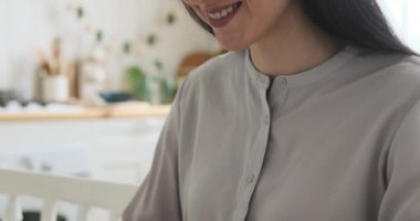 Mutfak masasında oturan Kafkas kökenli neşeli bir kadının yakın plan görüntüsü. Yüzünde ışıl ışıl bir gülümsemeyle, kendini dizüstü bilgisayara kaptırmış, verimliliğini gösteriyor..