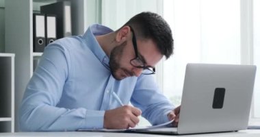 Modern ofisinde birden fazla iş yapan bir iş adamı. Dizüstü bilgisayarı ve kalemiyle, karmaşık grafikler ve çizelgeler içeren çeşitli belgeleri ustalıkla inceliyor..
