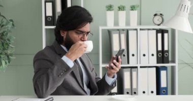 Hintli bir işadamı ve kravatlı bir takım elbiseli bir adam, bir molada masasında oturmuş, çayını yudumluyor ve hoş bir ifadeyle telefonunu karıştırıyor..