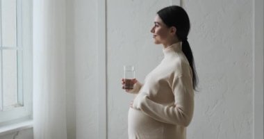 Hamile beyaz kadın pencerenin kenarında zarif bir şekilde duruyor ve ferahlatıcı su tadını şımartıyor. Nazik bir dokunuşla, büyüyen karnını okşar, annelik yolculuğunu kucaklar..