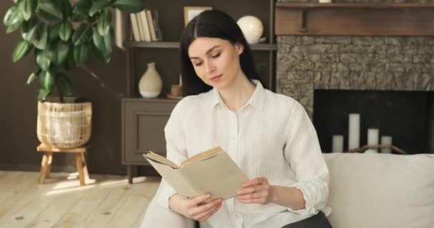 一个安静的白人女人坐在客厅的沙发上 全神贯注地看一本迷人的书 她平静的举止和专注的凝视显示出她对文坛的深深投入 — 图库视频影像