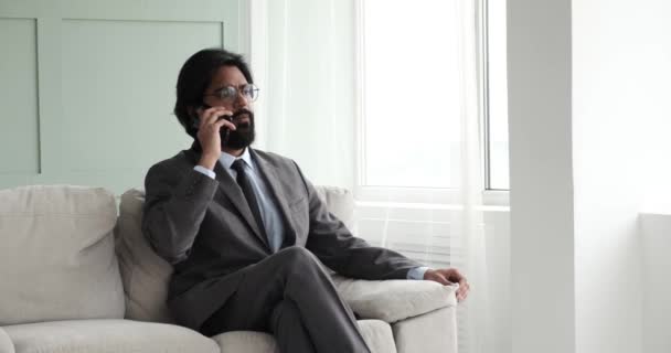 眼鏡をかけたインド人ビジネスマンとネクタイをしたスーツは 自宅のソファに自信を持って座って電話で話し合い 仕事について話し合っています 彼の集中し 決定的な表現は彼のプロ意識を伝える — ストック動画