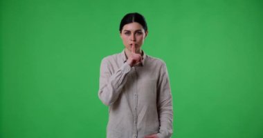 Tatmin olmamış beyaz bir kadın yeşil bir ekranda duruyor ve sus işaretini dudaklarının yanında gösteriyor. Sonra bir şeyi yasaklayarak parmağını ve başını sallıyor..