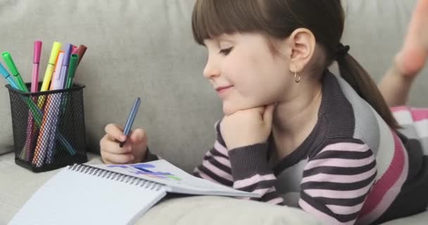 一个年龄在学龄前的年轻白人女孩躺在沙发上 她手里拿着笔 巧妙地 富有想象力地在笔记本上画了些特别的东西 — 图库视频影像