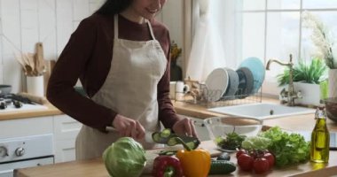 Kafkasyalı kadın mutfakta duruyor, günlük kıyafetler ve mutfak önlüğü giyiyor. Işıl ışıl bir gülümseme ile, hünerli bir şekilde taze bir salatalık hazırlar, ahşap bir kesme tahtasına keser..