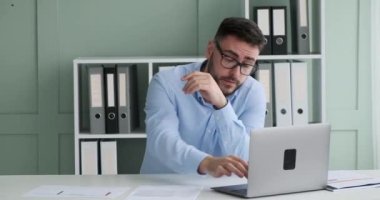 İş kıyafeti ve gözlük takan, sakallı, yorgun Avrupalı girişimci, ofisinde masasında oturuyor, belgeleri gözden geçiriyor ve yorgunluktan baş ağrısı çekiyor..