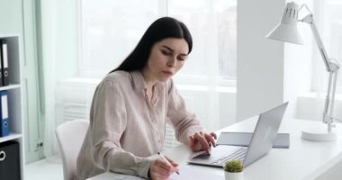 Ofisteki masasında çalışan gömlekli bir iş kadını. Yüzünde ciddi bir ifadeyle, belgelerine veri girmeye çalışıyor ve bunu laptopuyla karşılaştırıyor, bu da kafa karışıklığını gösteriyor..