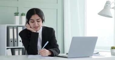 Siyah ceket ve beyaz gömlek giyen Hintli iş kadını ofisinde kulaklık takıyor. Müzik dinlerken, evrak işlerinde de dahil olmak üzere birden fazla işi sorunsuzca yapıyor..