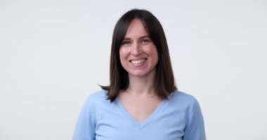 Güzel beyaz bir kadın beyaz bir arka planda duruyor, parlak, samimi bir gülümseme sergiliyor. Doğrudan kameraya bakarken dişleri görünür, pozitif ve neşe saçıyor..