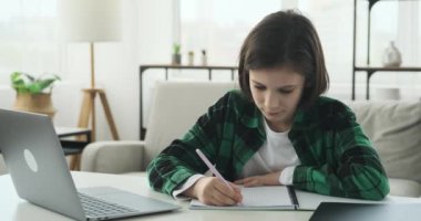 Öğrenci çocuk oturma odasındaki masada oturuyor. O aktif olarak dizüstü bilgisayar kullanarak online bir derse katılıyor. Yüzü ilgiyi yansıtıyor ve elleri deftere yazmakla meşgul..