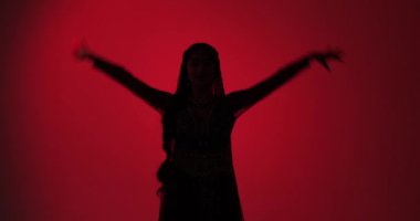 Genç Hintli dansçı, sari giyip muhteşem geleneksel bir dans sergiliyor. Kırmızı arka planda dans ediyor, zarif silueti Hint kültürünün güzelliğini ve zarafetini gösteriyor..