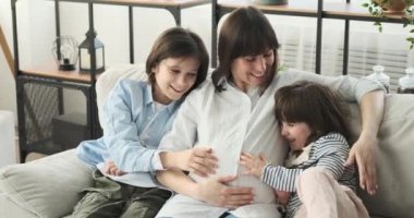 Oturma odasındaki rahat kanepede toplanan hamile bir anne, oğlu ve kızı. Şefkatli gülümsemeler ve dolup taşan sevgiyle, büyüyen bebek tümseğini sevgiyle kucaklarlar..