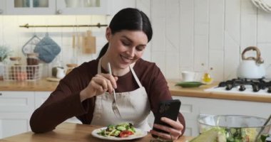 Neşeli beyaz kadın mutfak masasında oturmuş tabağından nefis bir salatanın tadını çıkarıyor. Geniş bir gülümseme ve bulaşıcı bir kahkaha ile telefonuna bakarken neşe ve pozitiflik yayıyor..